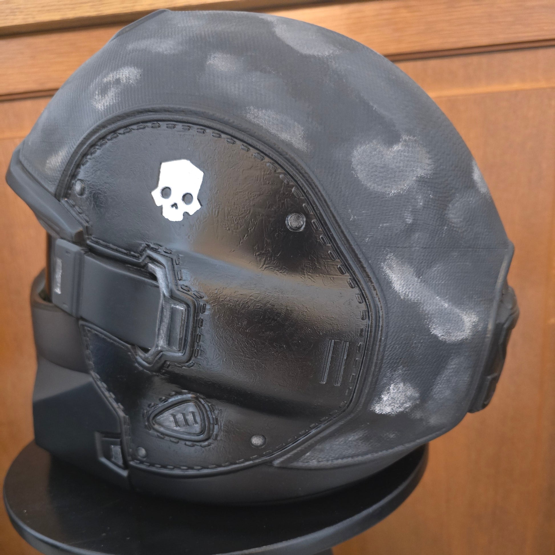 A black tactical helmet with a distressed gray camouflage pattern on the shell, featuring a glossy black visor and a white skull logo on the forehead, displayed on a stand against a wooden background.