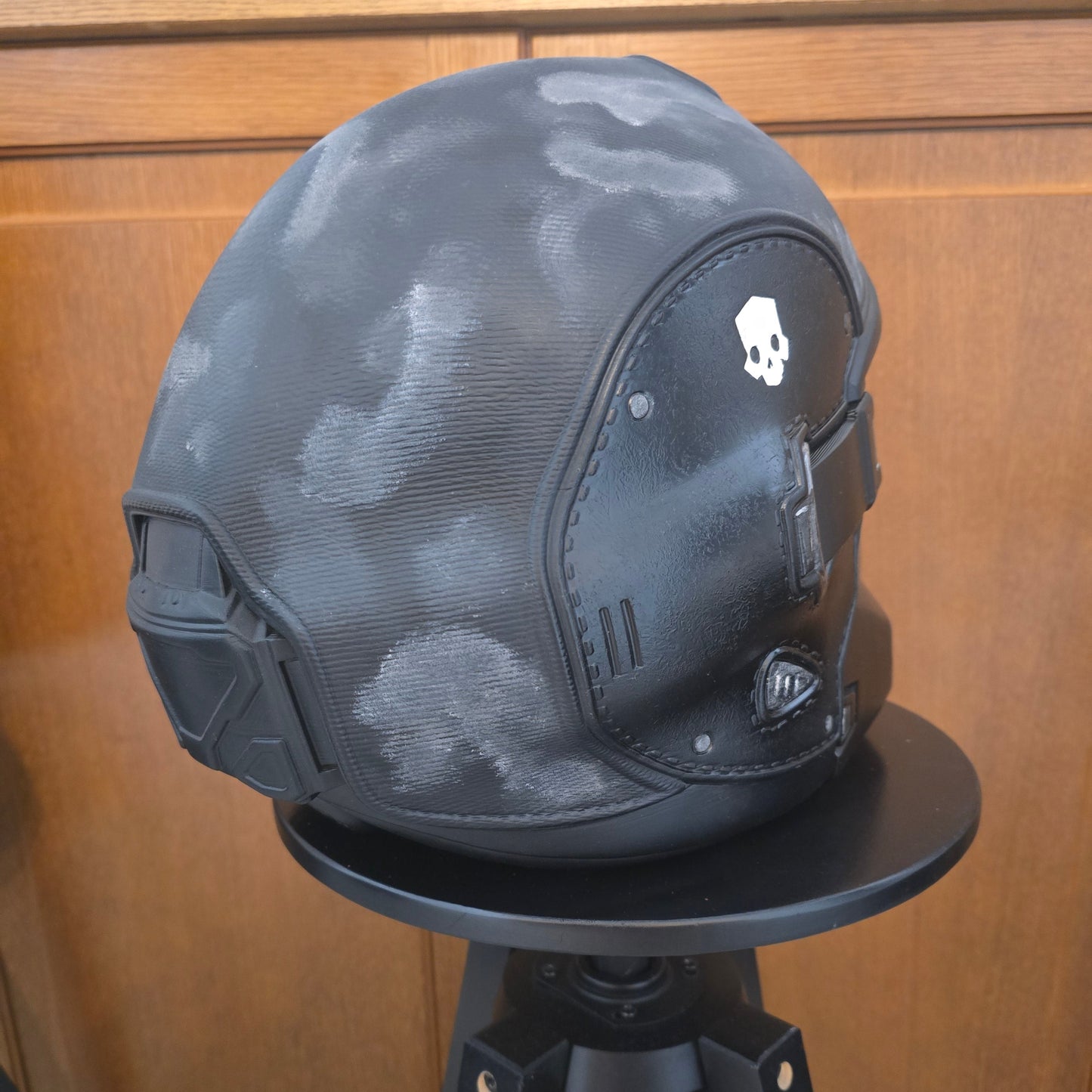 A black tactical helmet with a subtle gray camouflage pattern, displayed on a black stand against a wooden door background.
