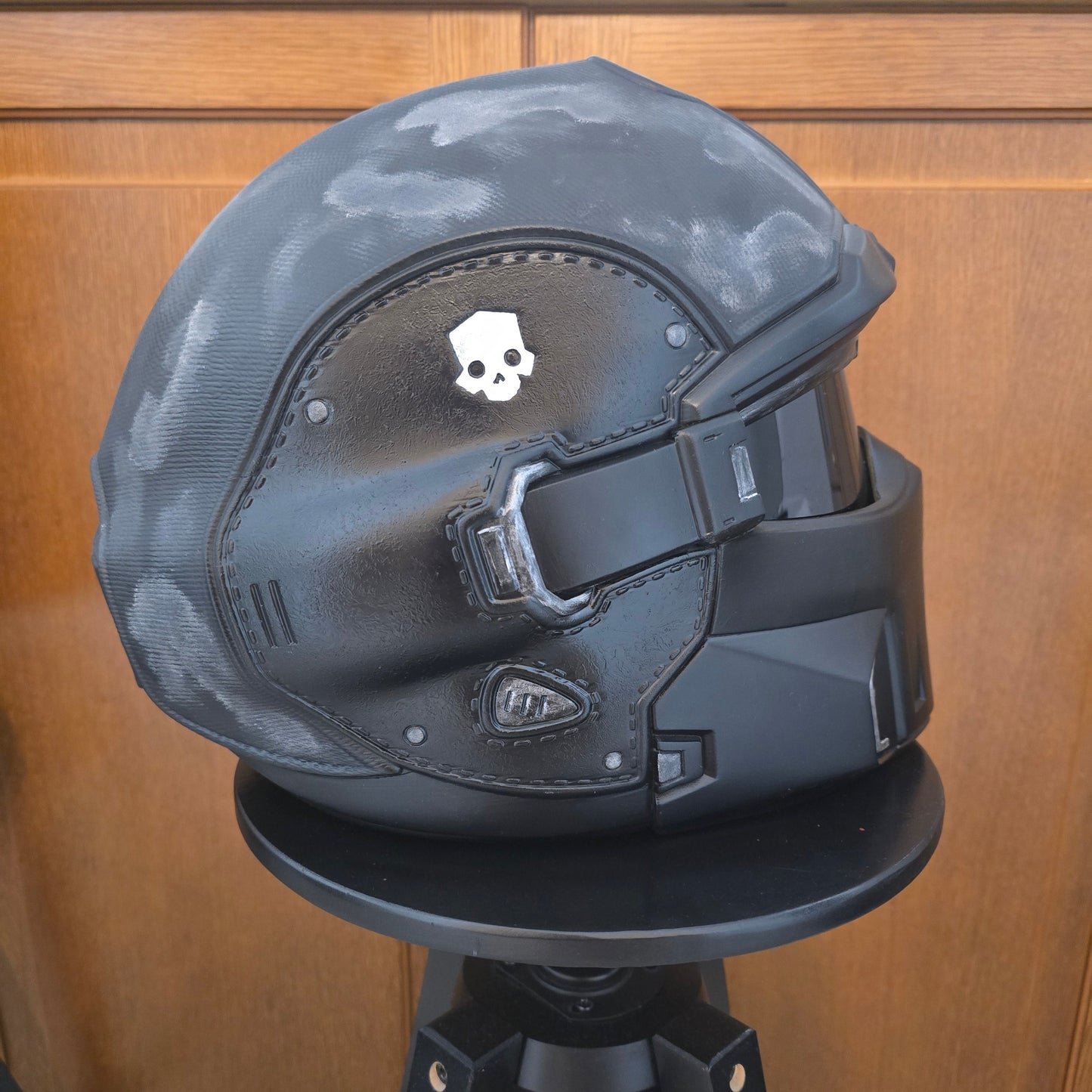 A black and dark gray futuristic helmet or headgear with molded panels and straps, photographed indoors against a wooden door background.