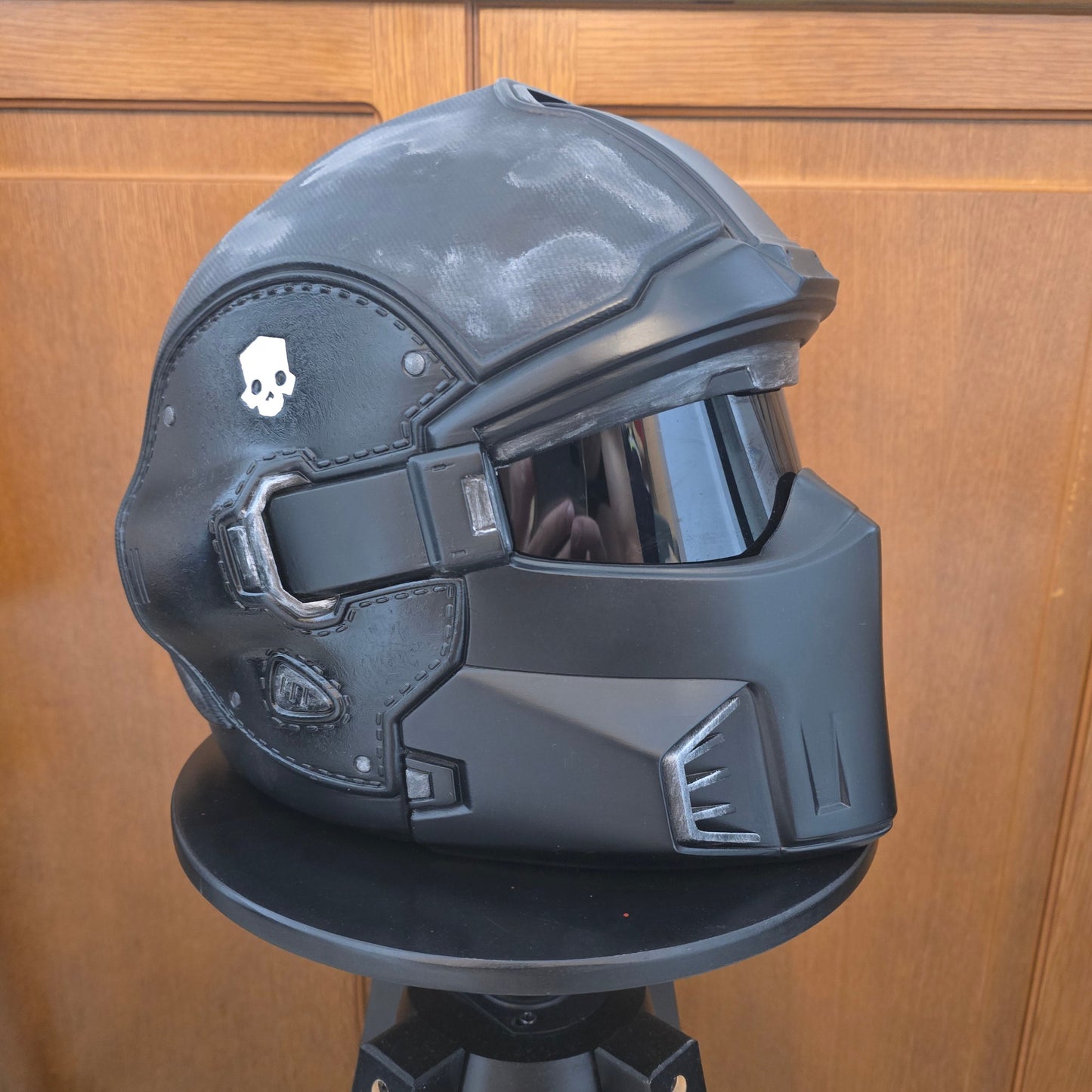 A black futuristic sci-fi helmet displayed on a black stand against a wooden door background.