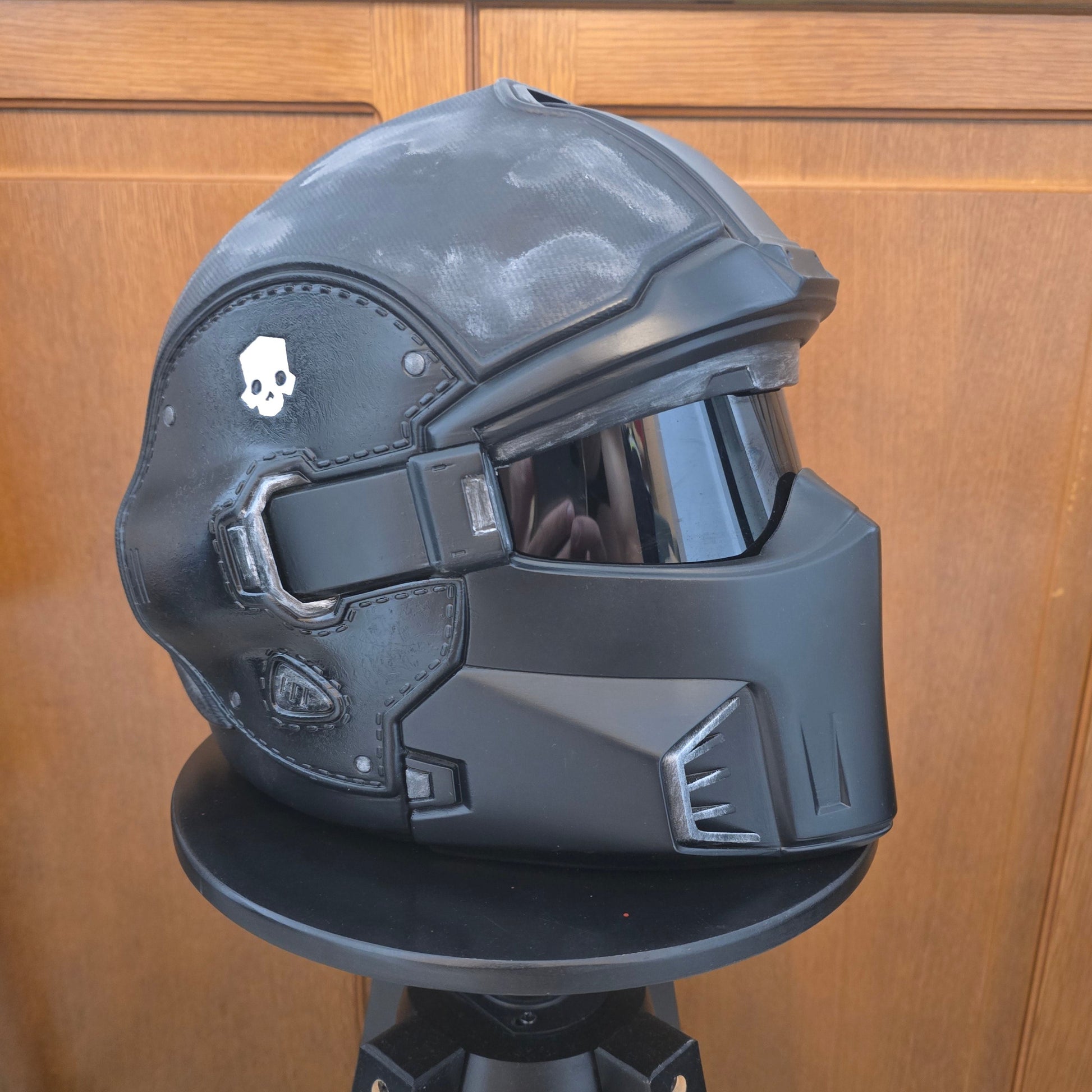 A black futuristic sci-fi helmet displayed on a black stand against a wooden door background.