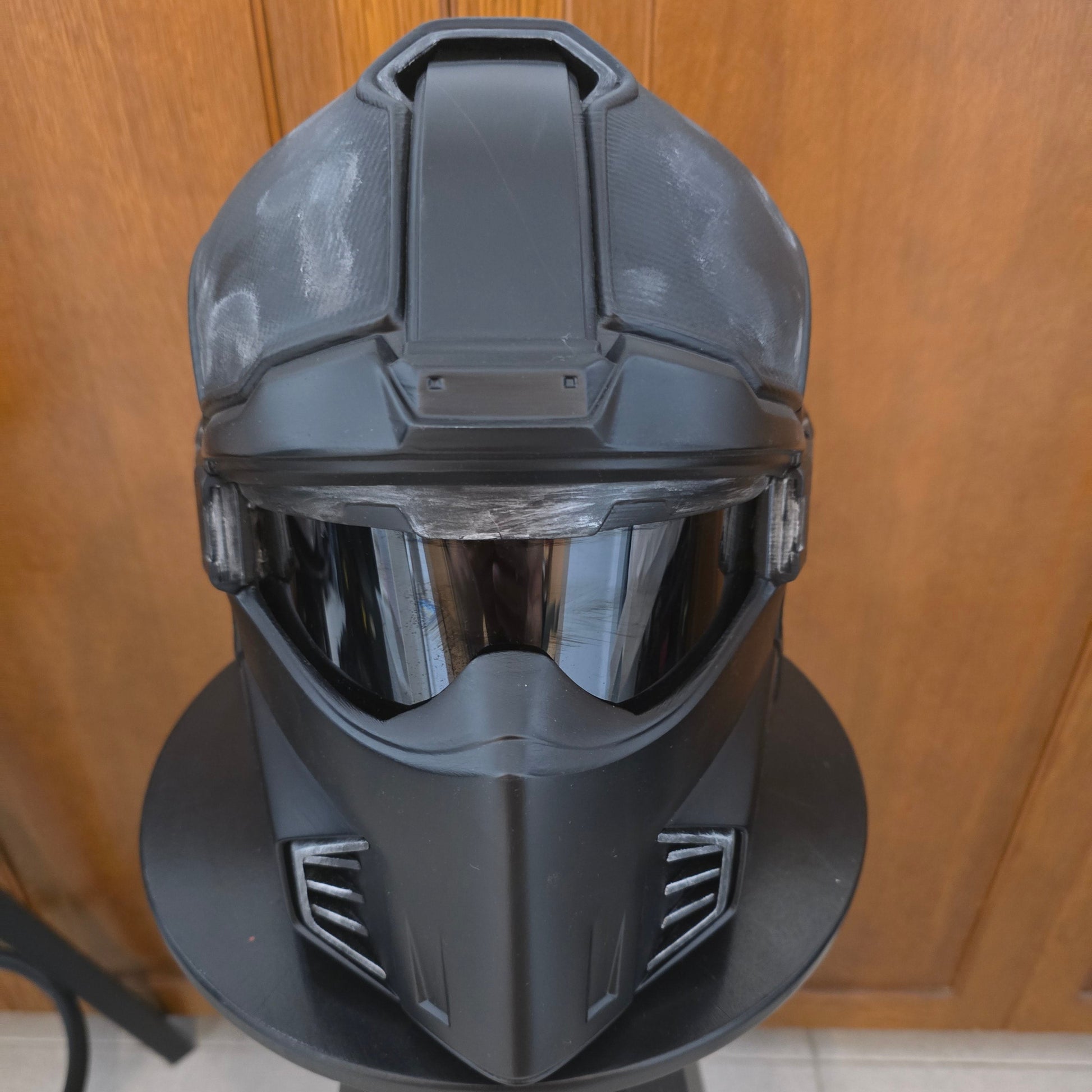 A black futuristic sci-fi helmet with a clear visor, displayed on a black stand in front of a wooden door.